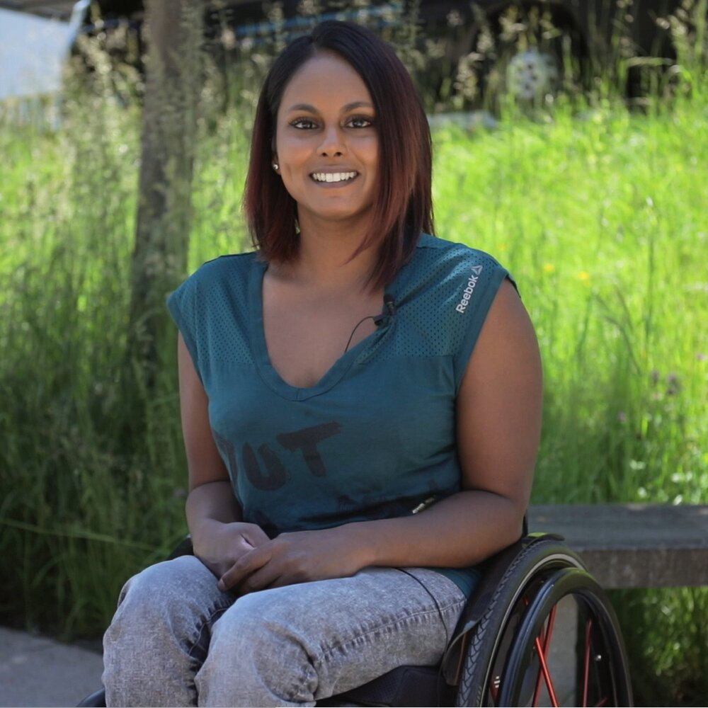 Alexandra Helbling hat dunkle, mittellange Haare und lächelt in die Kamera. Sie ist inkomplette Paraplegikerin und sitzt im Rollstuhl. Im Hintergrund ist eine grüne Wiese zu erkennen. | © Stiftung MyHandicap / EnableMe