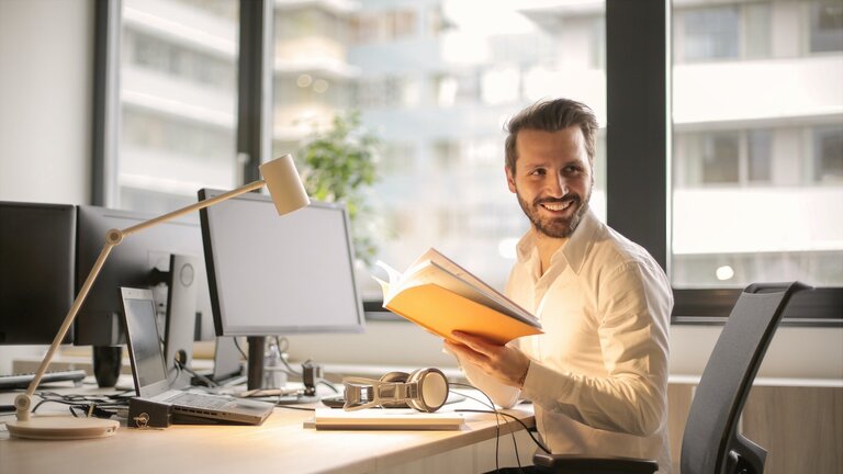 Ein Mann sitzt lachend am Bürotisch und hält ein aufgeschlagenes Heft in den Händen. | © pexels