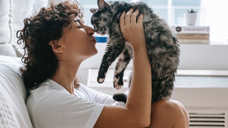 Bild einer jungen Frau mit lockigen Haaren, die eine grau-weisse Katze hochhält. Im Hintergrund sieht man nein Bett und ein Fenster. | © pexels