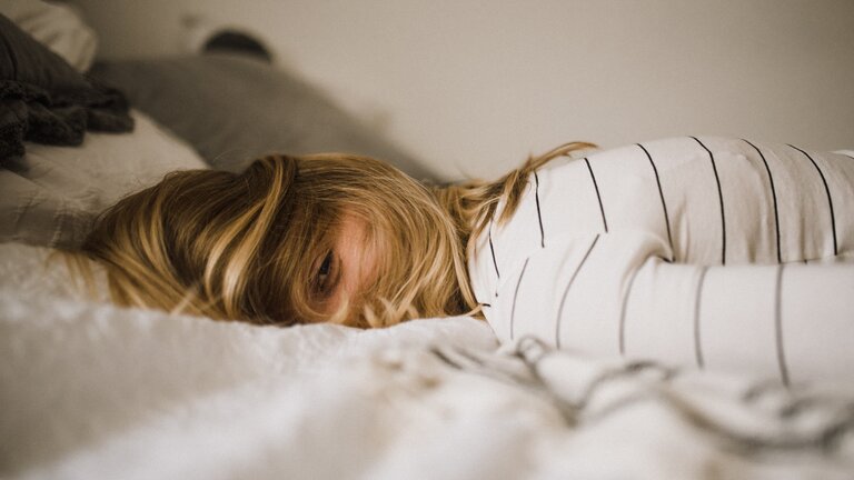 Nahaufnahme einer Frau, die auf einem Bett liegt. Sie trägt ein gestreiftes Oberteil, ihr Gesicht ist mit ihren blonden Haaren bedeckt. | © unsplash