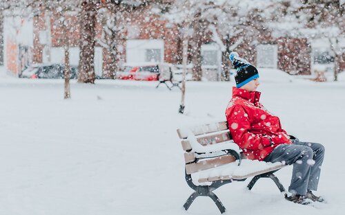 Ein Mann sitzt auf einer Bank in einer winterlichen Umgebung. Er trägt eine rote Jacke und eine blaue Pudelmütze. | © unsplash
