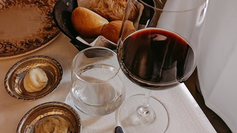 Ein Foto, auf dem ein Weinglas mit Rotwein und ein Brotkorb zu sehen sind. Im Hintergrund sieht man Besteck und einen Teller, der auf einem weissen Tischtuch steht. | © pexels