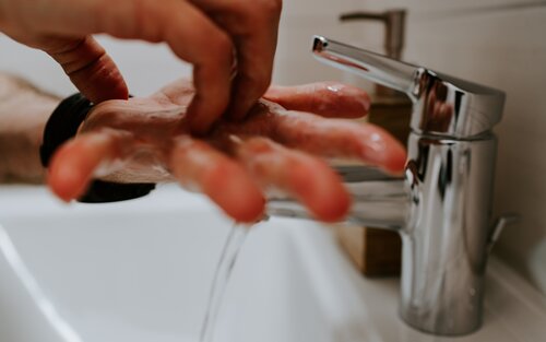 Zwei Hände werden unter fliessendem Wasser aus einem modernen, silbernen Wasserhahn gehalten. Es scheint, als würde sich die Person gründlich die Hände waschen. Der Fokus liegt auf den Händen und dem Wasserhahn, der Hintergrund ist unscharf. | © unsplash