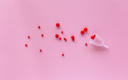 Das Bild zeigt einen rosa Hintergrund mit mehreren kleinen roten Pompons, die verstreut sind. In der rechten unteren Ecke befindet sich eine Menstruationstasse, die umgedreht ist und einige der roten Pompons enthält. | © pexels