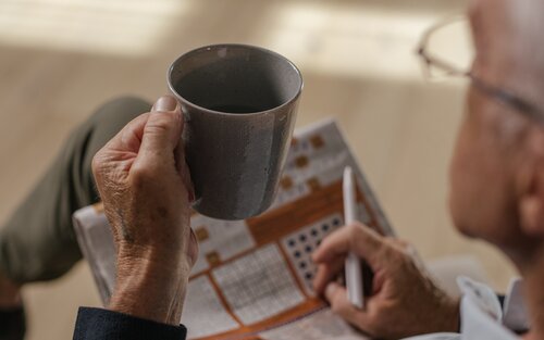 Das Bild eines älteren Mannes von hinten, in der Hand hält er eine Zeitung und eine Kaffeetasse. | © pexels