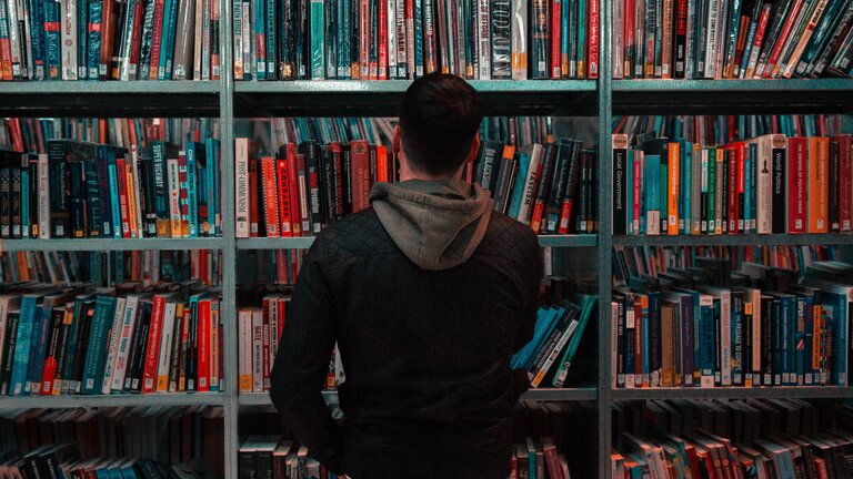 Ein Mann mit dunklen Haaren stent vor einem großen Bücherregal in einer Bibliothek. Er trägt eine Jeans und eine dunkle Jacke und steht mit dem Rücken zur Kamera. | © Unsplash