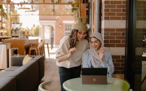 Zwei Frauen winken in die Webcam eines Laptops. Die eine steht hinten und trägt einen hellen Pulli, eine schwarze Hose und eine Mütze, die andere sitzt vorne und trägt eine hellblaue Bluse und ein helles Kopftuch. | © pexels