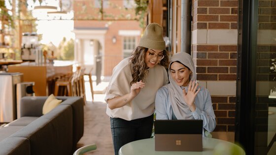 Zwei Frauen winken in die Webcam eines Laptops. Die eine steht hinten und trägt einen hellen Pulli, eine schwarze Hose und eine Mütze, die andere sitzt vorne und trägt eine hellblaue Bluse und ein helles Kopftuch. | © Geeignete Berufe für Menschen mit Depressionen