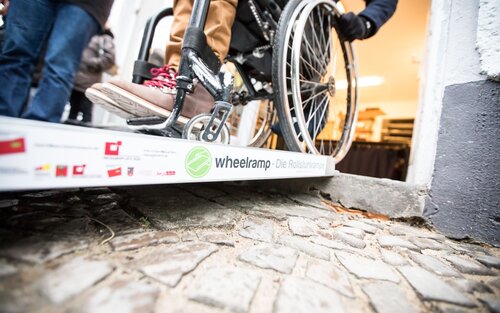 Nahaufnahme eines Rollstuhls, der über eine mobile Rampe fährt. | © Gesellschaftsbilder