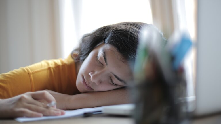 Eine junge Frau sitzt an ihrem Arbeitstisch, auf dem ein Notizbuch, ihr Laptop und ein Behälter mit Stiften stehen. Die Frau hat ihren Kopf auf ihren Arm und den Tisch gelegt und schläft. | © pexels