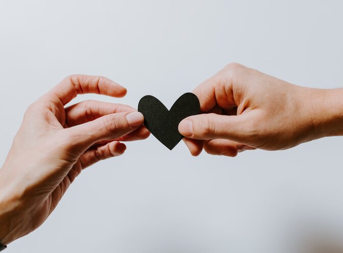 Zwei Hände, die sich ein schwarzes Herz aus Papier übergeben. | © Unsplash / Kelly Sikkema