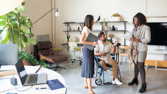 Ein dreiköpfiges Team von Personen mit und ohne Behinderungen bei einer informellen Stehsitzung im Büro. | © Pexels / Kampus Production