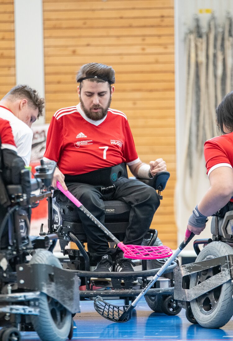 Eine Gruppe von Sportler:innen im Rollstuhl, die sich mit ihren Schlägern nach dem Ball strecken. Alle haben rote Trikots an, gespielt wird in der Halle.