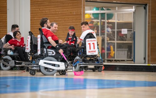 Das Team mit rot-weissen Leibchen spielt Powerchair-Hockey im Rollstuhl. Sie befinden sich in einer Halle. | © PCH Worldcup