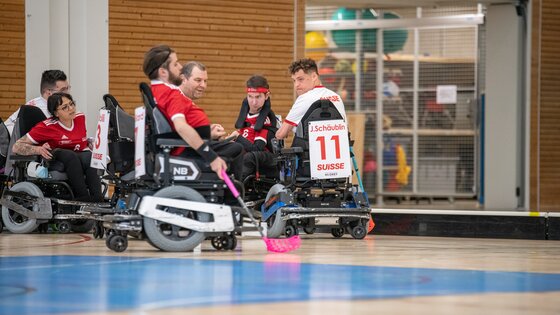Das Team mit rot-weissen Leibchen spielt Powerchair-Hockey im Rollstuhl. Sie befinden sich in einer Halle. | © PCH Worldcup