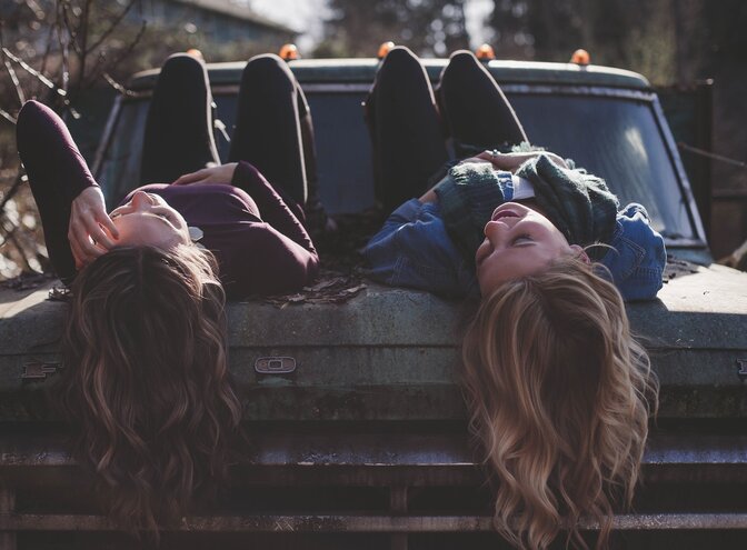 Foto von zwei jungen Frauen, die auf dem Rücken auf einem Auto liegen, sich unterhalten und lachen. | © pixabay
