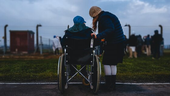 Person im Rollstuhl mit unterstützender Person vor Zaun | © Unsplash