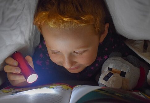 Foto eines Jungen, der mit einer Taschenlampe unter der Bettdecke ein Buch liest. | © pexels