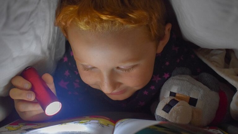 Foto eines Jungen, der mit einer Taschenlampe unter der Bettdecke ein Buch liest. | © pexels
