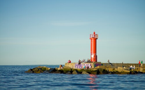 Ein roter Leuchtturm neben dem Meer mit Personen auf den Felsen um ihn herum verstreut | © unsplash