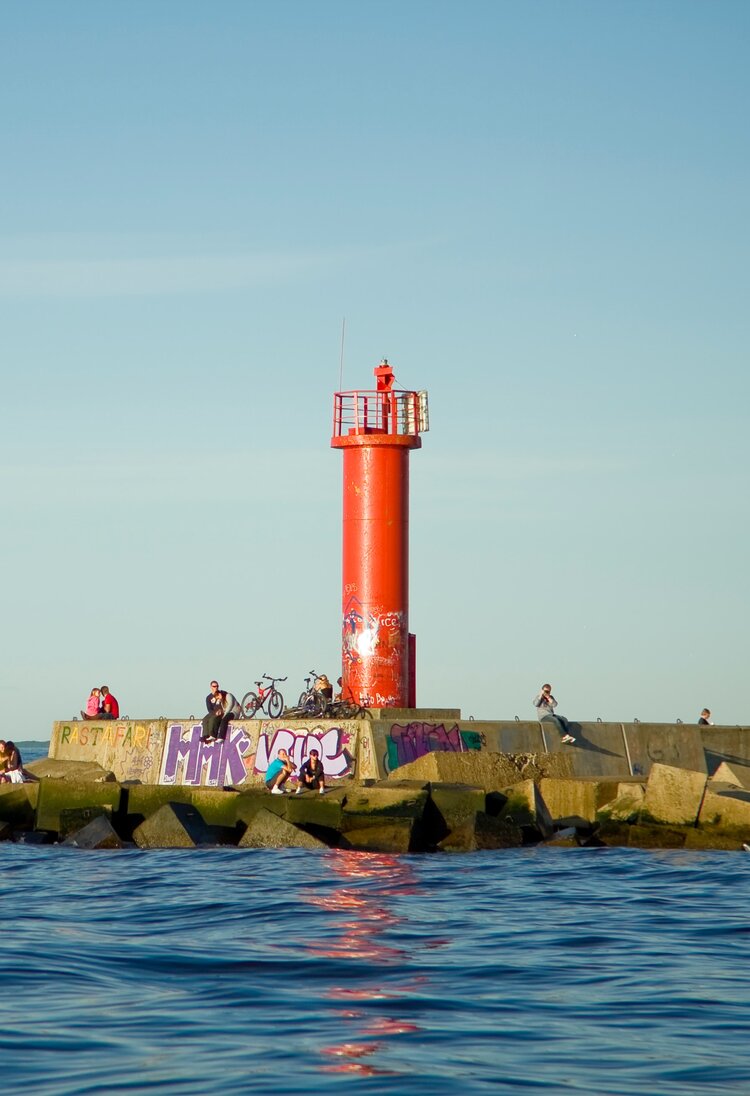 Ein roter Leuchtturm neben dem Meer mit Personen auf den Felsen um ihn herum verstreut | © Unsplash