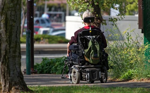 Foto von einer älteren Person, die mit ihrem Elektrorollstuhl einen Gang entlangfährt. Die Person ist nur von hinten zu sehen. | © pexels