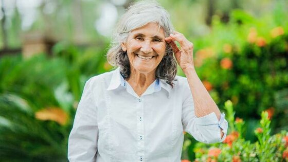 Eine Seniorin mit weißer Bluse steht lächelnd in einem Garten. | © pexels