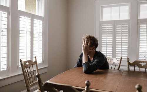 Ein Mann sitzt an einem Holztisch, hat den Kopf in die Hände geschützt und sieht traurig aus. | © pexels