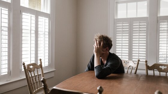 Ein Mann sitzt an einem Holztisch, hat den Kopf in die Hände geschützt und sieht traurig aus.