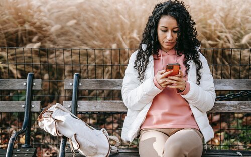 Eine Frau mit schwarzen Locken und einer weissen Jacke sitzt auf einer Holzbank und blickt auf das Smartphone in ihrer Hand. | © pexels
