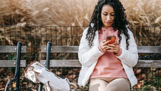Eine Frau mit schwarzen Locken und einer weissen Jacke sitzt auf einer Holzbank und blickt auf das Smartphone in ihrer Hand. | © pexels
