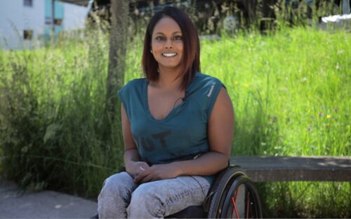 Alexandra Helbling hat dunkle, mittellange Haare und lächelt in die Kamera. Sie ist inkomplette Paraplegikerin und sitzt im Rollstuhl. Im Hintergrund ist eine grüne Wiese zu erkennen. | © Stiftung MyHandicap / EnableMe
