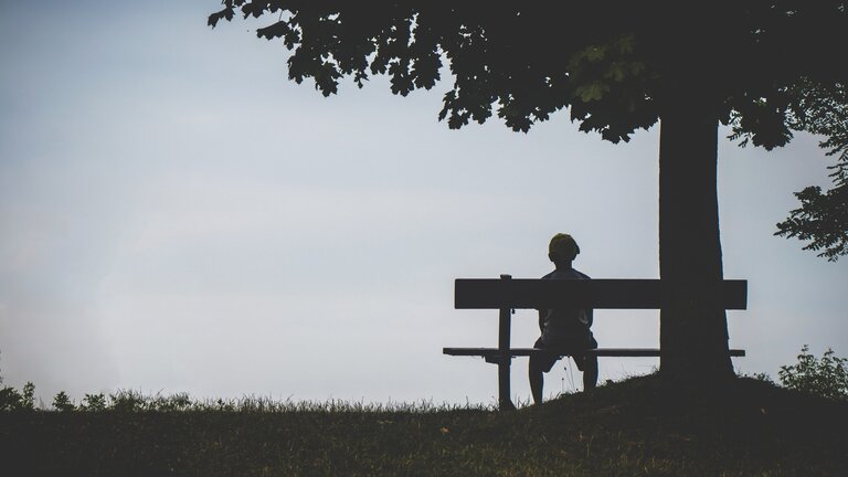 Junge sitzt alleine auf einer Bank, neben ihm ein Baum. | © pexels