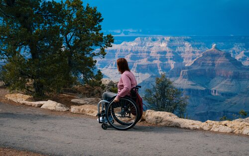 Sur la photo, on voit une jeune femme assise dans un fauteuil roulant. Elle regarde vers le bas une vallée.