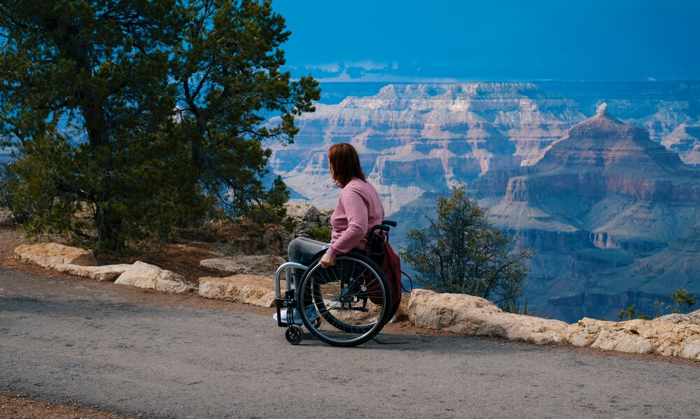 Auf dem Bild ist eine junge Frau zu sehen, die im Rollstuhl sitzt. Sie schaut auf ein Tal hinab. | © unsplash