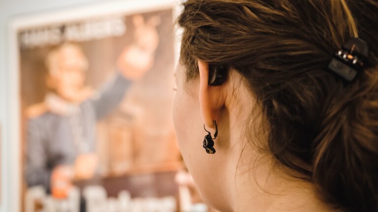 Une jeune femme avec un appareil auditif regarde une affiche floue. | © Gesellschaftsbilder.de
