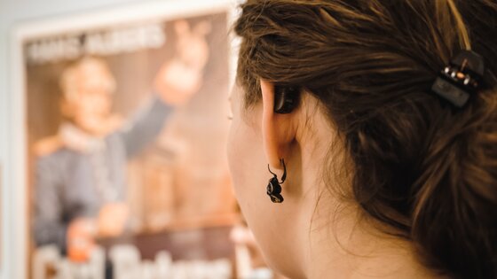 Junge Frau von hinten mit Hörgerät sieht auf ein verschwommenes Plakat. | © Andi Weiland/ Gesellschaftsbilder.de