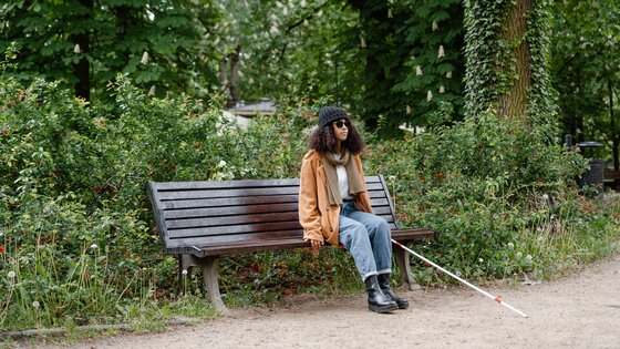 Eine Frau mit Blindenstock und Sonnenbrille sitzt auf einer Parkbank. Im Hintergrund sind Bäume und viel Grün zu erkennen. | © Pexels/Mart Production