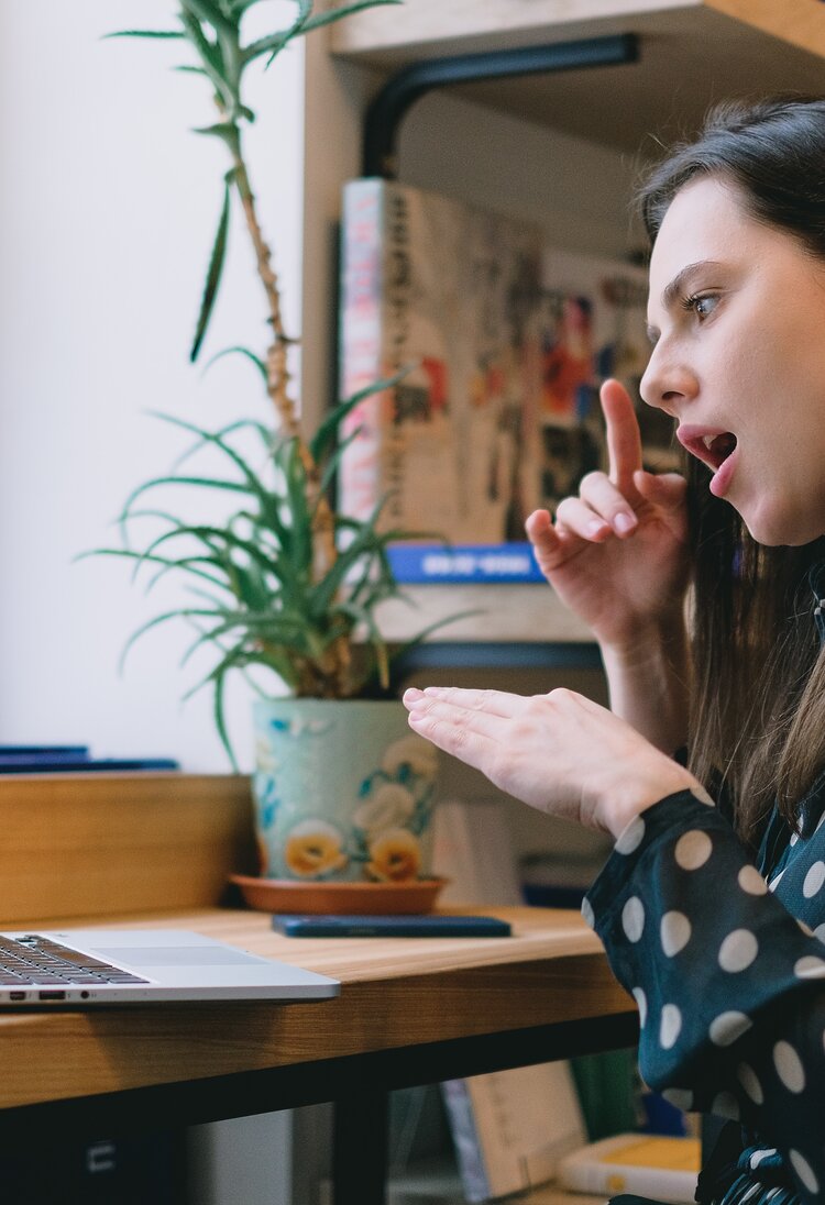 Eine Frau sitzt vor einem Laptop und spricht in Gebärdensprache. Sie trägt eine gepunktete Bluse, im Hintergrund ist ein Bücherregal zu sehen. | © Pexels / SHVETS production