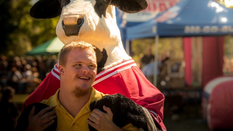 Mensch mit Trisomie 21 wird von hinten von einer Stoff-Kuh umarmt. | © unsplash