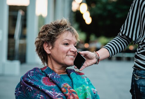 Un assistant tient un téléphone portable à l'oreille d'une femme handicapée. | © Andi Weiland | Gesellschaftsbilder.de