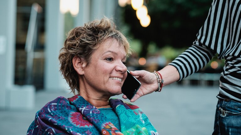 Assistenzperson hält einer Frau mit einer Behinderung das Handy ans Ohr. | © Andi Weiland | Gesellschaftsbilder.de