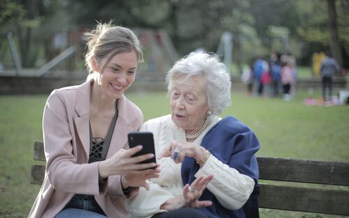 Eine jüngere Frau zeigt einer Seniorin etwas auf ihrem Handy, beide lächeln. | © pexels