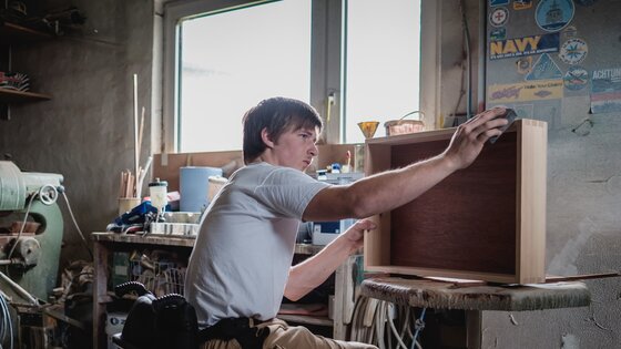Mann sitzt im Rollstuhl in einer Werkstatt und schleift ein Holzmöbel. | © Gesellschaftsbilder.de