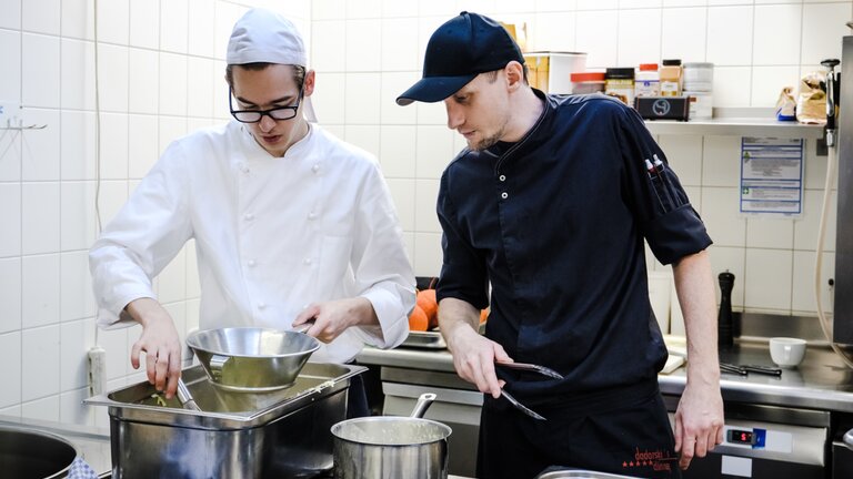 Ein junger Koch benützt Küchenutensilien während sein Ausbildner zuschaut. | © Gesellschaftsbilder.de