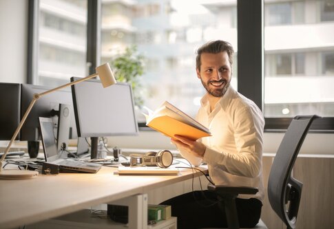 Ein Mann sitzt lachend am Bürotisch und hält ein aufgeschlagenes Heft in den Händen. | © pexels