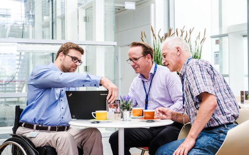 Drei Arbeitskollegen diskutieren an einem Tisch, wobei einer davon mobilitätseingeschränkt ist. | © Gesellschaftsbilder.de