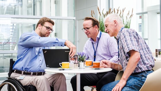 Drei Arbeitskollegen diskutieren an einem Tisch, wobei einer davon mobilitätseingeschränkt ist. | © Gesellschaftsbilder.de