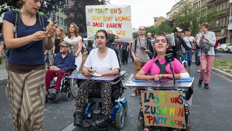 Menschen mit und ohne Rollstuhl sind auf einer Strasse versammelt. An einem Rollstuhl ist ein Plakat befestigt: «Lasst uns zusammen Party machen» | © Gesellschaftsbilder.de