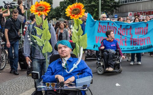 Menschenmenge ist auf Strasse versammelt mit Banner "behindert und verrückt feiern". Zuvorderst ist eine Person im Rollstuhl mit blauen Haaren und zwei Sonnenblumen am Rollstuhl befestigt. | © Gesellschaftsbilder.de